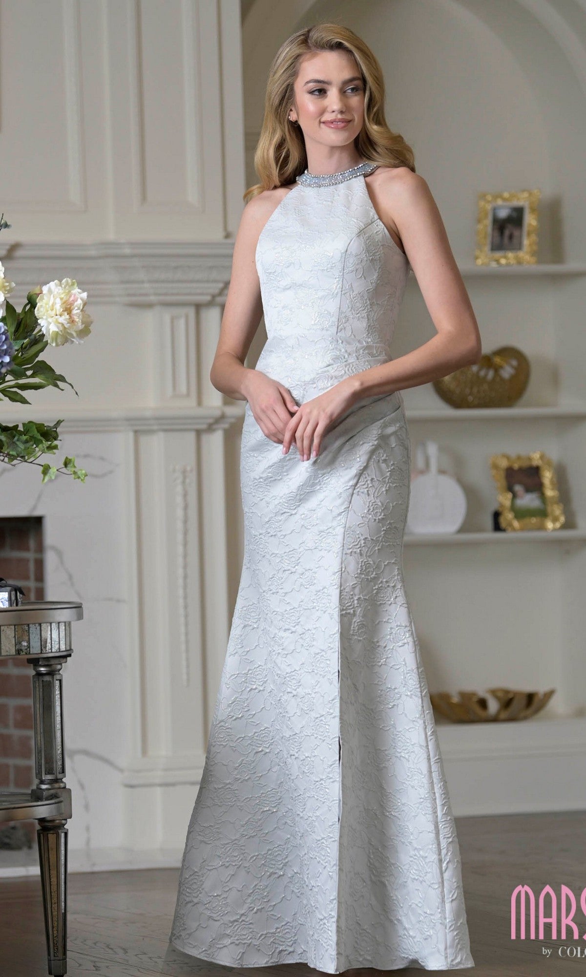 A woman in a sleeveless, floor-length white Marsoni M356 formal dress with intricate lace stands indoors by a fireplace and decorative shelves.